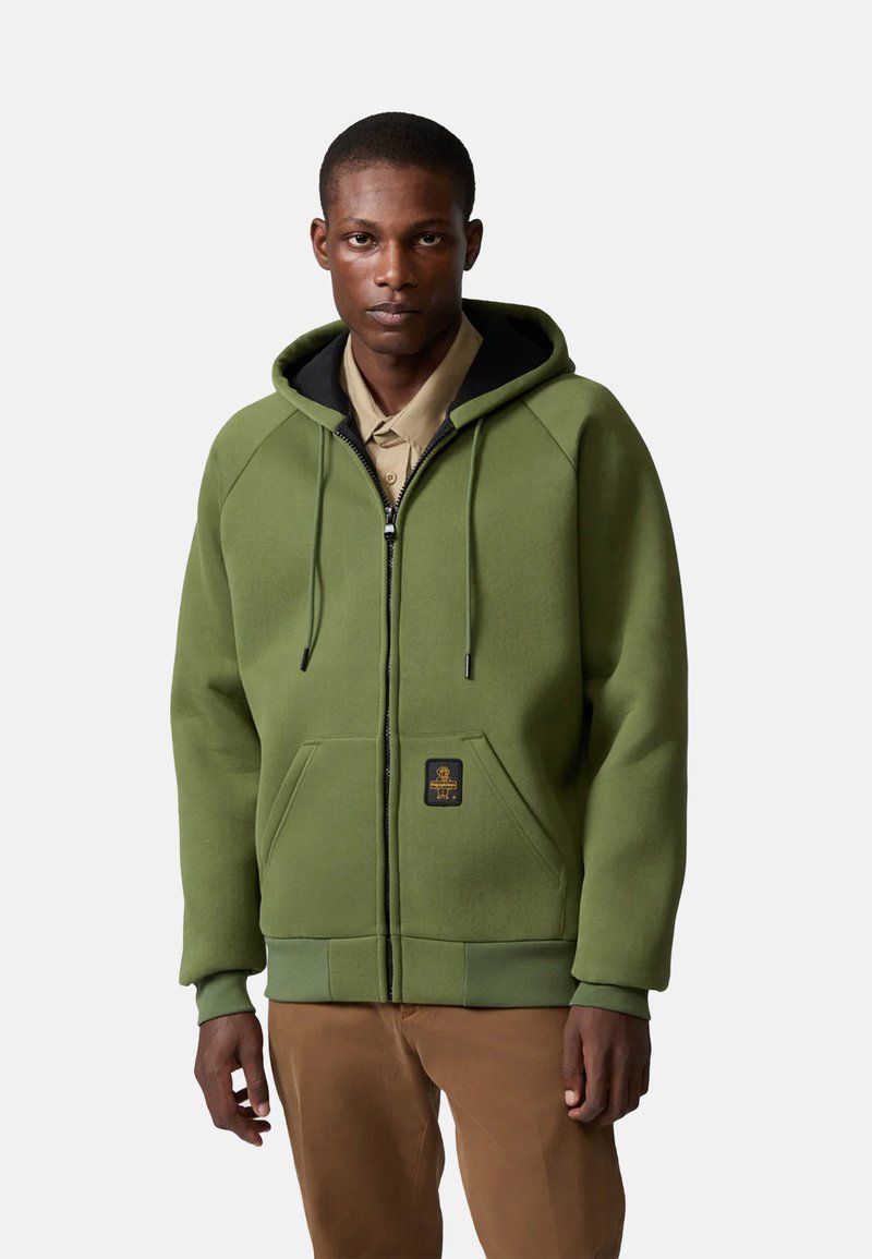 Felpa verde con zip e cappuccio interno nero, tasche frontali, polsini a costine e patch con logo sul petto. Texture morbida simile al pile.