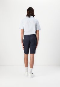 Vue arrière d'une personne portant une chemise à manches courtes bleu clair et un short marine foncé, associé à des baskets blanches et des chaussettes gris clair.