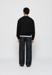Mann med mørkt krøllete hår iført svart genser, hvit T-skjorte, løse mørkeblå jeans og beige sko som står med ryggen til på hvit bakgrunn.