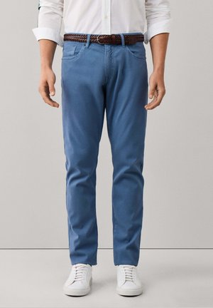 Person trägt ein weißes Hemd, blaue Hosen mit einem braun geflochtenen Gürtel und weiße Sneaker und steht vor einem schlichten Hintergrund.