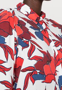 Collared blouse featuring a white background with large red and navy floral patterns, textured fabric, and visible button details.