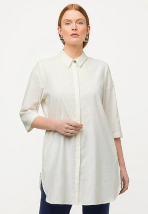 Camisa blanca de botones con cuello, mangas tres cuartos y aberturas laterales. Hecha de una tela ligera, con un único botón en el cuello.