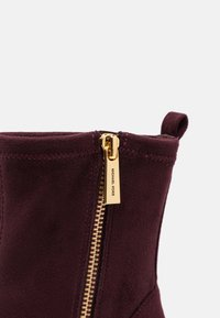 Gros plan sur une botte en daim bordeaux avec une tirette de fermeture éclair métallique dorée gravée "MICHAEL KORS" et une languette en tissu en haut.