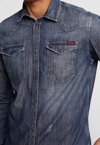 Camicia in denim blu scuro con un design aderente, due tasche sul petto, bottoni a pressione e dettagli di usura discreti. Colletto classico con accenti di cucitura.