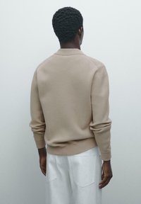 Homme avec des cheveux courts et frisés portant un pull beige à manches longues et un pantalon blanc, se tenant face à un fond gris clair uni.