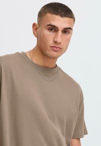 Camiseta de algodón marrón con cuello redondo, mangas cortas y una textura suave, combinada con un sencillo collar de cadena de plata.