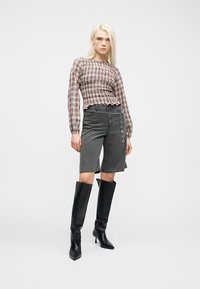 Νεαρή γυναίκα που φοράει ένα καρό τοπ με μακριά μανίκια, ψηλόμεση γκρι βερμούδα μέχρι το γόνατο με ζώνη αλυσίδα και μαύρες μπότες μέχρι το γόνατο με τακούνι, όρθια.