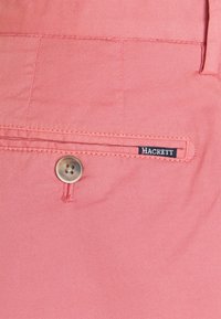 Růžové bavlněné šortky s hladkou texturou, s kapsou na knoflík a malou značkovou štítkem "Hackett" poblíž kapsy.