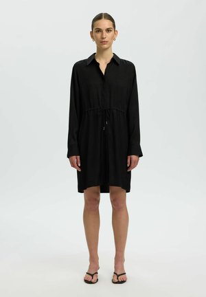 Femme debout portant une robe chemise noire à manches longues avec col et ceinture à la taille, associée à des sandales à talons noires sur un fond uni.