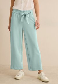 Pantalons larges bleu clair à taille nouée, en tissu doux ; présentent une texture lisse et un ourlet cousu. Associés à des baskets blanches.