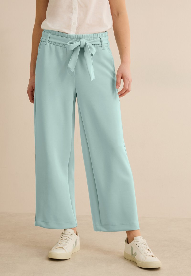 Pantalons larges bleu clair à taille nouée, en tissu doux ; présentent une texture lisse et un ourlet cousu. Associés à des baskets blanches.