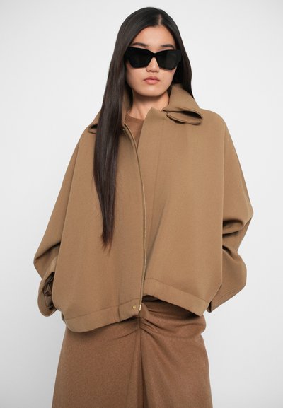 Smeđa, oversized jakna s visokim ovratnikom i patentnim zatvaračem, izrađena od teksturiranog materijala. Nosena preko usklađenog smeđeg outfita i stilizirana s naočalama.