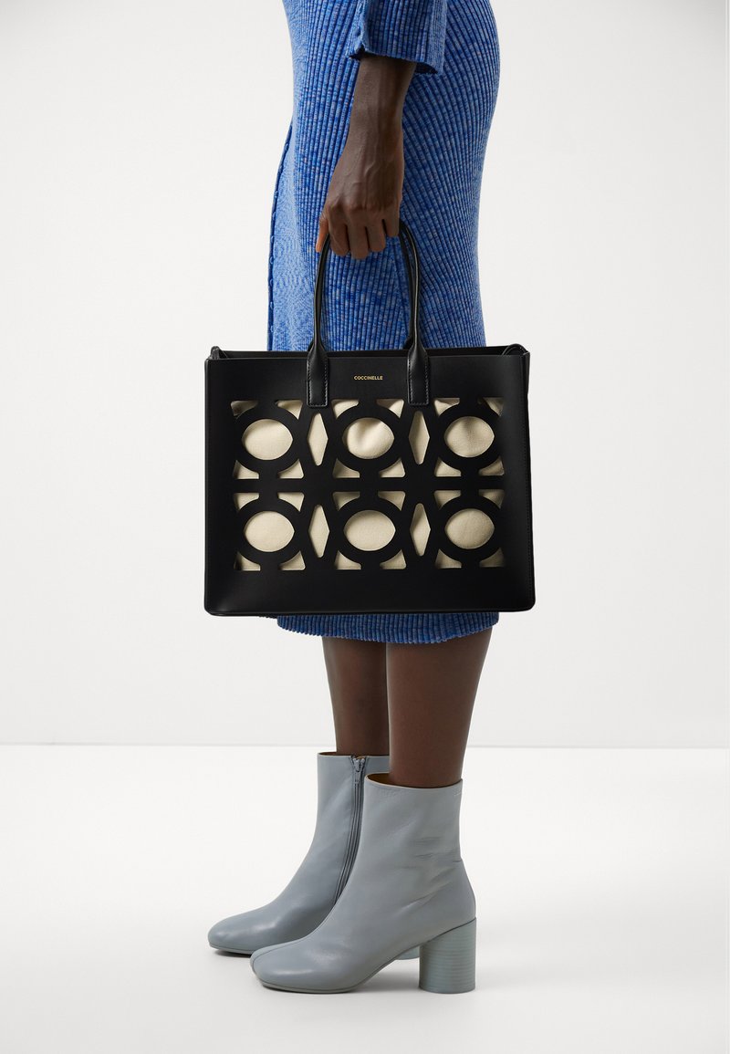 Sac fourre-tout en cuir noir avec des motifs circulaires découpés révélant un intérieur clair, doté de deux poignées et d'un logo sur le devant.