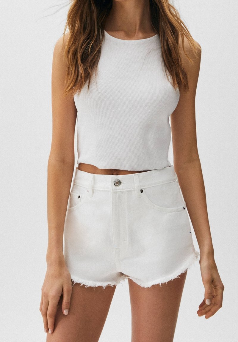 Femme portant un crop top sans manches blanc et un short en denim blanc taille haute avec des ourlets effilochés, se tenant devant un fond uni.