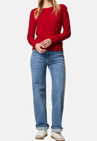 Pull rouge texturé à manches longues, associé à un jean large bleu clair et des baskets blanches, avec des poignets retroussés et une coupe décontractée.