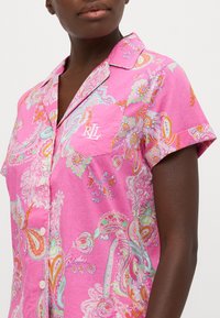 Rozā rakstaina pidžamu krekls ar apkakles izgriezumu, īsām piedurknēm, priekšējo kabatu ar izšūtām iniciāļiem un pogu aizdari.