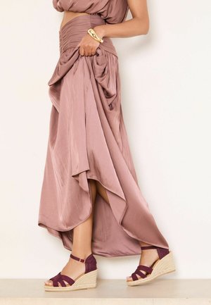 Femeie purtând o fustă mov cu volane și sandale mov cu platformă și curele la gleznă, ridicând ușor tivul fustei, stând într-un fundal pal.