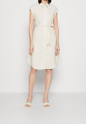 Vrouw draagt een beige overhemdjurk met korte mouwen, ceintuur en knopen, en gevlochten sandalen met open teen, staand tegen een effen achtergrond.