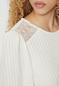Blusa bianca texturizzata con strisce verticali e dettaglio in pizzo sulle spalle. Il tessuto è leggero con un aspetto morbido e rigato.