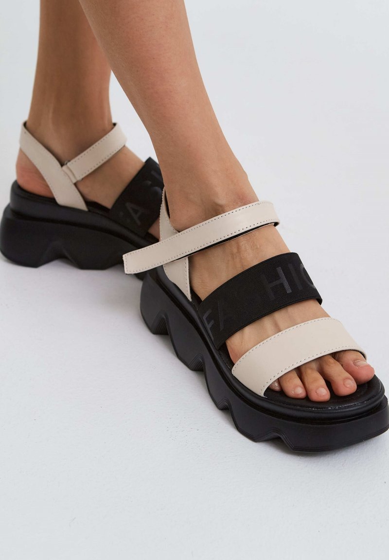 Gros plan de pieds portant des sandales à plateforme épaisses beige et noire avec des semelles épaisses et des brides réglables sur un fond clair.