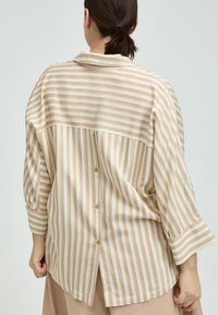 Woman with dark hair in a low ponytail wearing a beige and white striped buttoned blouse, viewed from behind with hands relaxed at sides.