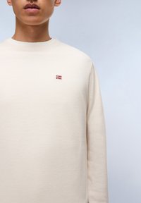 Sudadera beige claro hecha de tejido texturizado, con cuello redondo y un pequeño emblema de bandera bordado en el pecho.