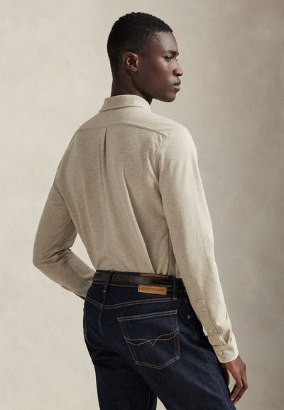 Camisola bege de manga comprida com gola, usada com jeans de ganga azul escuro com detalhes de costura e um remendo de couro na cintura.