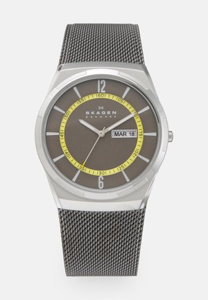 Silver Skagen wristwatch with brown dial, yellow minute ring, mesh strap, date display showing Mar 18, and silver hour and second hands.