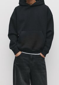 Černá mikina s kapucí a přední kapsou, vyrobená z měkké tkaniny. Má žebrované manžety a lem, doplněná tmavými džínami.