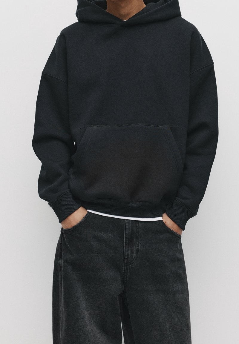 Černá mikina s kapucí a přední kapsou, vyrobená z měkké tkaniny. Má žebrované manžety a lem, doplněná tmavými džínami.