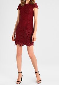 Žena v krátkých rukávech, burgundském krajkovém šatech se zvlněným lemem, v kombinaci s černými lodičkami na anklet, stojící na bílém pozadí.