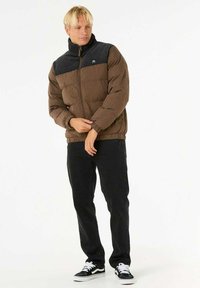 Bruine pufferjack met zwarte kraag, ritssluiting en elastische manchetten. Gecombineerd met zwarte jeans en zwarte sneakers. Eenvoudig ontwerp, warme textuur.