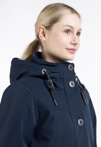 Giacca con cappuccio blu navy in tessuto testurizzato, con bottoni frontali e cordini. Presenta un colletto alto e un design pulito e minimalista.
