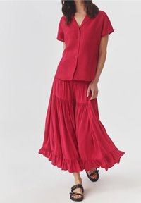 Rode knoopjesblouse met korte mouwen, gecombineerd met een vloeiende, enkellange rode rok met ruffles aan de onderkant en comfortabele sandalen.