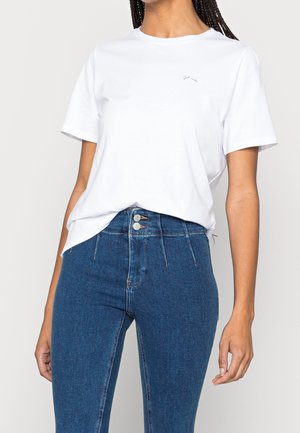 Vrouw draagt een eenvoudige witte korte mouwen T-shirt, gestopt in een hoogtailored blauwe spijkerbroek met dubbele knoopsluiting.