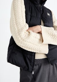 Black puffer vest with a high collar and front zipper, paired with a chunky cream cable-knit sweater featuring ribbed cuffs.