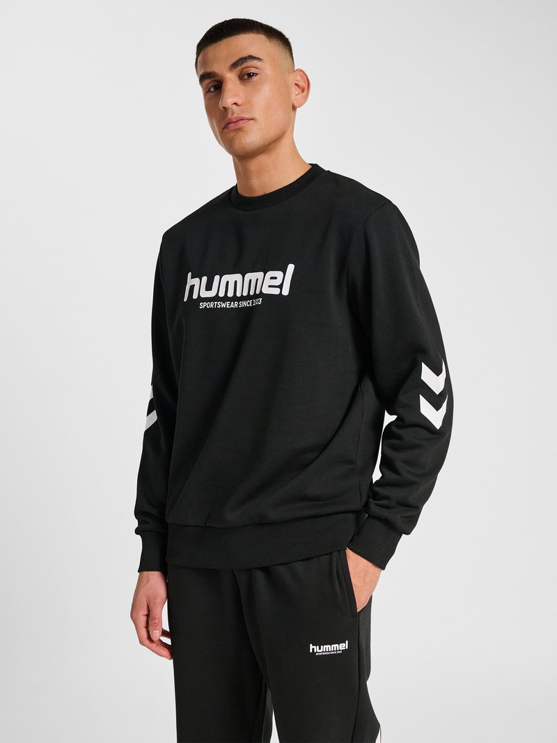 Junger Mann mit kurzen Haaren, der einen schwarzen hummel-Sweatshirt und passende schwarze hummel-Hosen trägt, mit einer Hand in der Tasche vor einfarbigem Hintergrund stehend.
