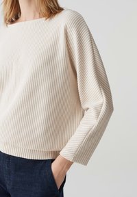 Top beige chiaro con strisce verticali testurizzate, scollatura larga e maniche lunghe, abbinato a pantaloni di jeans scuri. Finitura in tessuto morbido.