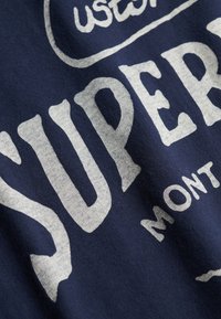 Gros plan sur un tissu bleu marine avec un grand texte blanc "SUPER" et un texte plus petit "MONT" dans un style vintage et usé.