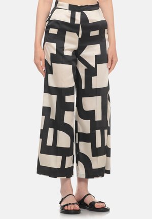 Donna che indossa pantaloni a vita alta a gamba larga con motivo geometrico nero e beige e sandali neri aperti in punta, in piedi su uno sfondo semplice.