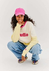Ležérní outfit zahrnující krémovou a žlutou mikinu s nápisem "utto" v červené, světle modré baggy džíny a růžovou čepici. Outfitu dominuje textura tenisek.