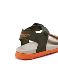 Sandale vert olive avec une semelle orange, dotée d'une sangle en Velcro, d'une semelle intérieure texturée et d'une marque orange sur la sangle.