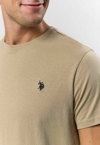 Beige tričko s kulatým výstřihem vyrobené z hladkého bavlněného materiálu, s malým vyšitým logem v námořnické modři na levé straně hrudi. Krátké rukávy.