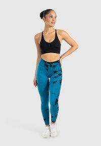 Kobieta stoi z ręką na biodrze, ubrana w czarny stanik sportowy, niebiesko-czarne legginsy tie-dye i białe sneakersy na jednolitym tle.