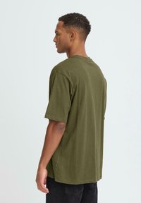 Tricou din bumbac de culoare verde măsliniu, cu mâneci scurte, guler rotund și o croială lejeră. Fanta laterală la tiv și detaliu discret cu etichetă de branding.