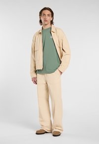 Lys beige jakke, grøn t-shirt og matchende beige bukser. Jakken har to franske lommer. Brune sko med strukturerede såler.