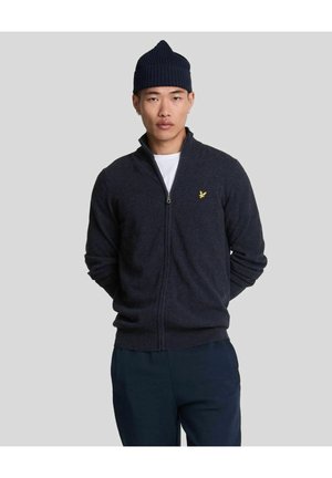 Mann trägt einen dunkelblauen Reißverschlusspullover mit einem kleinen gelben Emblem, dunkelblaue Hose, weißes Unterhemd und eine dunkelblaue Strickmütze und steht vor einem grauen Hintergrund.