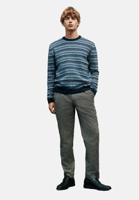 Blauer gemusterter Pullover mit Rundhalsausschnitt, graue strukturierte Hose und schwarze Schuhe. Das Model steht mit den Händen in den Taschen vor einem schlichten Hintergrund.