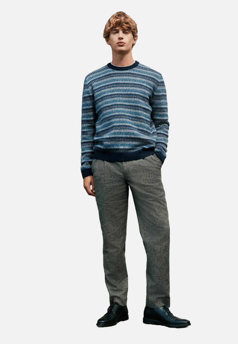 Blauer gemusterter Pullover mit Rundhalsausschnitt, graue strukturierte Hose und schwarze Schuhe. Das Model steht mit den Händen in den Taschen vor einem schlichten Hintergrund.