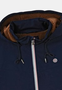 Marinblå jacka med brunt fleecefoder, har dragkedja med röda och vita detaljer, snörning och en cirkulär logotyp.